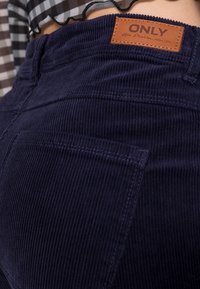 Pantalons en velours côtelé navy avec une finition texturée, comportant une poche arrière et une étiquette en cuir marron portant la mention "ONLY" avec des détails de couture.