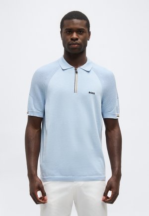 Férfi világoskék, rövid ujjú, cipzáras BOSS logós galléros pólót visel, fehér nadrággal, egyszínű háttér előtt áll.