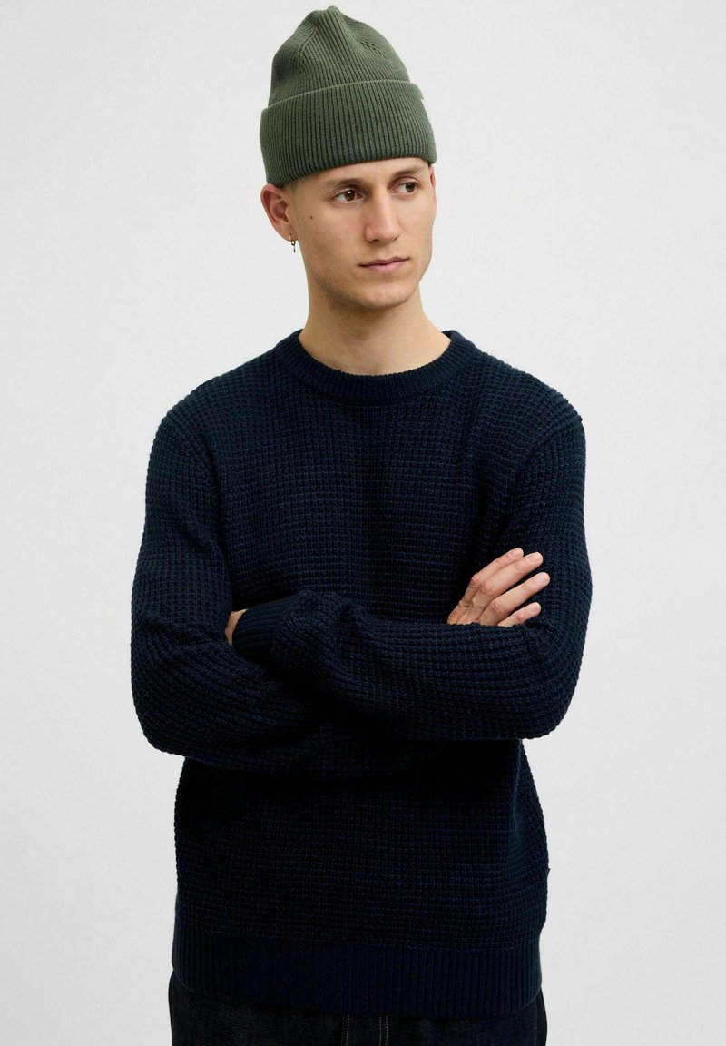 Mariniers gebreide trui met een textuurpatroon en ribbelmanchetten, gedragen met een groene beanie. Het model staat met gekruiste armen tegen een effen achtergrond.
