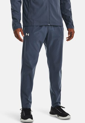 Mann trägt dunkelblaue Sporthose mit Under Armour-Logo, schwarze Sneakers mit weißen Sohlen und passende dunkelblaue Jacke.