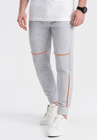 Graue Sweatpants aus weichem Material mit orangefarbenen Akzenten entlang der Nähte und gerippter Struktur an den Knieplatten; kombiniert mit weißen Sneakers.