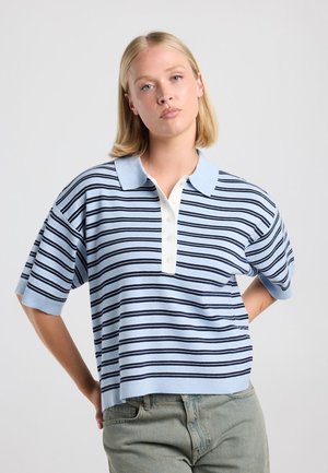 Young woman with short blonde hair wearing a light blue and navy striped short-sleeve polo shirt and gray jeans, standing with one hand on hip.