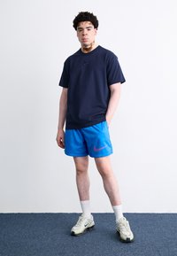 Mornarsko modra bombažna majica s Nike logotipom, v kombinaciji z modrimi kratkimi hlačami z roza Nike oznako in svetlo sivimi športnimi čevlji z črnimi akcenti.