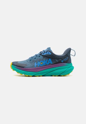 Zapatilla de running HOKA con parte superior de malla en azul y gris, media suela en verde y morado, suela exterior texturizada y tacos de tracción amarillos.