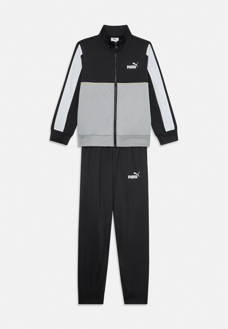 Melnas un pelēkas treniņtērps ar iešūtu jaku ar augstu apkakli un baltām akcentiem, apvienots ar atbilstošām melnām biksēm, abiem ar logotipu.