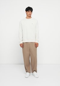 Lys grå genser med ribbestrikkede mansjetter, kombinert med løse beige bukser. Modellen står i hvite sneakers mot en ensfarget bakgrunn.