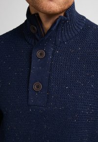 Pull en maille bleu marine avec un col côtelé, deux fermetures à boutons marron et un motif texturé avec des éclats de couleurs plus claires.