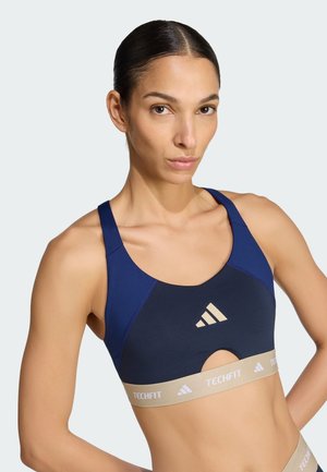 Atletische vrouw met donker haar in een blauw en beige Adidas Techfit sportbeha, staand met één hand op de heup tegen een witte achtergrond.