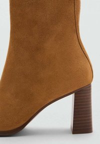 Stivale alla caviglia in suede marrone con punta quadrata e tacco in legno massiccio e rigato. La tomaia è liscia, con una texture delicata e cuciture pulite.