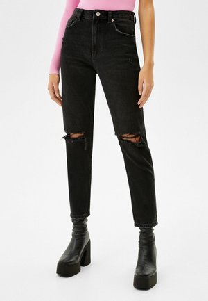 Femme portant un jean noir déchiré taille haute, des bottes noires à plateforme et à talons, et un haut rose à manches longues côtelé, debout devant un fond blanc.