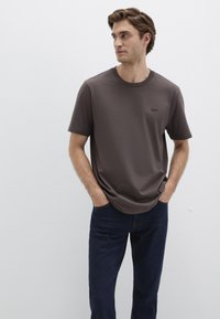 Kurzärmliges, dunkelbraunes T-Shirt mit rundem Ausschnitt und kleinem Logo auf der Brust, kombiniert mit dunkelblauen Jeans. Glatte Textur.