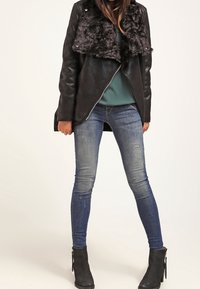 Chaqueta negra de cuero sintético con cuello de piel, llevada sobre una blusa verde azulado, combinada con jeans skiny azul desgastados y botas negras de tobillo.