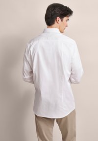 Weißes Baumwollhemd mit klassischem Kragen, langen Ärmeln und abgerundetem Saum, kombiniert mit beigen Hosen.