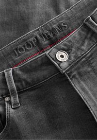 Vaqueros de mezclilla gris oscuro con una superficie texturizada. Presenta una etiqueta bordada "JOOP JEANS", costuras rojas y un botón de metal plateado.