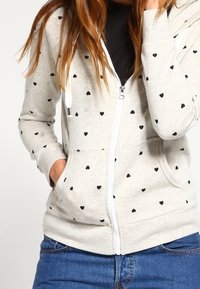 Sudadera con capucha y cremallera en gris claro con patrones de corazones negros, hecha de tela suave. Cuenta con capucha, bolsillos delanteros y puños elásticos.