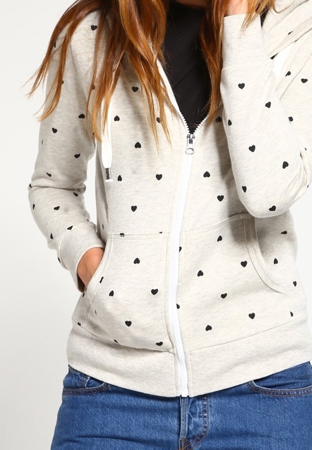 Sudadera con capucha y cremallera en gris claro con patrones de corazones negros, hecha de tela suave. Cuenta con capucha, bolsillos delanteros y puños elásticos.