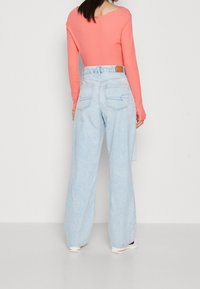 Top de manga larga en color coral combinado con unos vaqueros de pierna ancha de cintura alta en azul claro. Los vaqueros cuentan con dos bolsillos traseros y un parche de cuero marrón en la cintura.