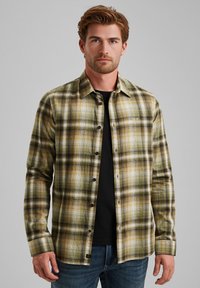 Camicia di flanella a quadri in tonalità verdi, gialle e nere; design con bottoni; maniche lunghe; indossata su una t-shirt nera; con cuciture visibili.