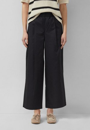 Osoba ubrana w luźne czarne spodnie z szerokimi nogawkami, beżowy sweter w czarne paski oraz beżowe sznurowane buty, stojąca na tle gładkiej szarej ściany.