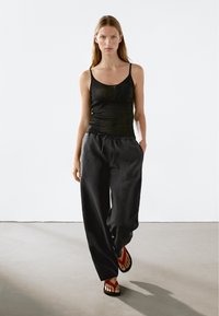 Schwarzes geripptes Tanktop und lose schwarze Hosen mit einem elastischen Bund, ergänzt durch rote Flip-Flops, alles vor einem neutralen Hintergrund.