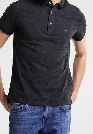 Tummanharmaa poolopaita, jossa on klassinen kaulukset, kolmen napin lista ja pieni logo rinnassa, yhdistettynä sinisiin farkkuihin.