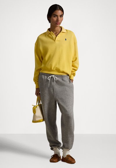 Kobieta ubrana w żółtą bluzkę polo z długim rękawem, szare spodnie dresowe, białe skarpetki i brązowe mokasyny, trzyma beżowo-żółtą torebkę, stoi na białym tle.