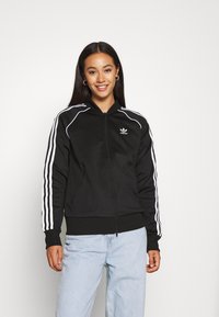 Sudadera con cremallera negra con rayas blancas en las mangas y logo. Cuenta con un alto cuello y dos bolsillos frontales, hecha de una tela suave.