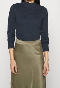 Marinblå långärmad topp med texturerat tyg och volangkrage, kombinerad med en satängkjol i olivgrönt, halvåtsittande silhuett, slät yta.