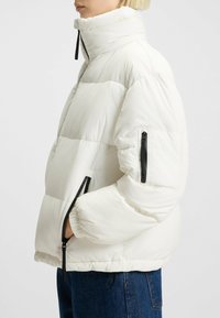 Vit puffjacka med hög krage, synliga sömmar och en zip-ficka på ärmen. Mjuk textur, elastiska muddar och nederkant.