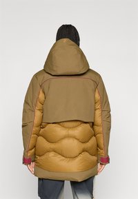 Pufjakke i brune nuancer, med hætte, en quiltet tekstur og kontrasterende bordeaux syninger. Designet inkluderer et lagdelt rygdetalje.