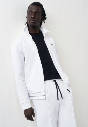 Mand iført hvid lynlåstrøje og bukser med sort skjorte nedenunder, stående mod en lysegrå baggrund, kigger til siden.