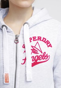 Sudadera con cremallera de color gris claro, con el logotipo bordado en rosa "Superdry Angels", cordones blancos, una tela texturizada y una cremallera plateada.