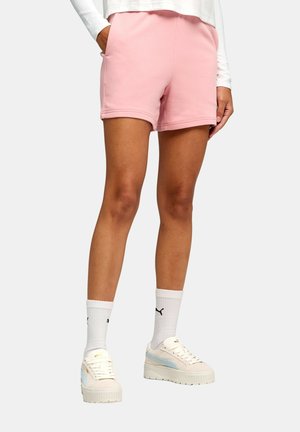 Persona che indossa pantaloncini rosa, maglia bianca a maniche lunghe, calzini bianchi Puma e sneakers beige con plateau, in piedi davanti a uno sfondo uniforme.