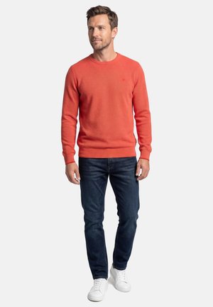 Man wearing orange long-sleeve sweater, dark blue jeans, and white sneakers, standing against a plain light background.