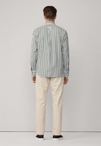 Camisa de botones a rayas en verde y blanco, con un cuello alto, llevada con pantalones beiges y zapatos oscuros. Aparece en un fondo de configuración neutra.