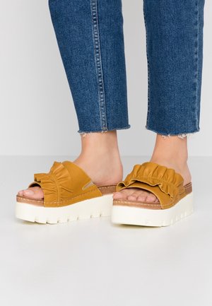 Gele slip-on sandalen met gerimpelde bovenkant en witte plateauzool. Getextureerd materiaal en open teen ontwerp zijn te zien. Spijkerbroek met rafelige zoom zichtbaar.