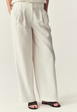 Bredbenede hvide linnedbukser båret med sorte åbne sandaler, hænder delvist i lommerne, stående på neutral baggrund.