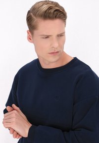 Sötétkék pulóver, puha anyagból készült, bordázott nyakkal és mandzsettával, a mellkasán diszkrét, dombornyomott logóval.