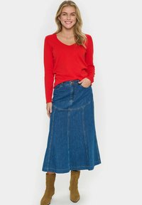 Pull rouge à col en V et manches longues associé à une jupe maxi en jean bleu avec une légère évasement et des détails cousus. Porté avec des bottines beige.