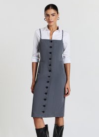 Graues knielanges Hemdkleid aus strukturierter Stoff, mit dünnen Trägern, vorderen Taschen und schwarzen Knöpfen. Wird über einem weißen Hemd getragen.