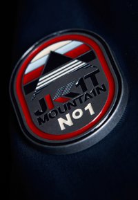 Badge rond avec une bordure rouge. Le design central présente un motif de montagne, des accents métalliques noirs et argentés, et le texte "JK1T MOUNTAIN No1."