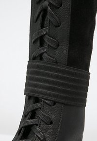 Černá bota vyrobená z hladké kůže a měkkého semiše. Obsahuje texturovaný pásek kolem kotníku a tkané tkaničky pro nastavení.