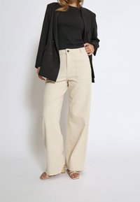 Femme portant un blazer noir et un top avec un pantalon beige taille haute à jambes larges et des sandales à talons ouvertes, debout devant un fond uni.