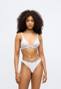 Λευκό σετ εσωρούχων που περιλαμβάνει ένα τρίγωνο μπραλέτ με ρυθμιζόμενες τιράντες και αντίστοιχο θόνγ, και τα δύο με ελαστική ζώνη με λογότυπο.