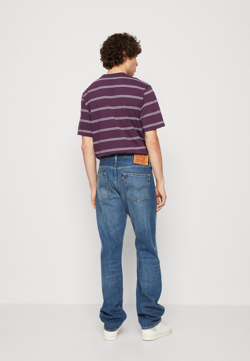 Homme de dos portant un t-shirt rayé violet, un jean bleu avec une étiquette en cuir et des baskets blanches sur un fond uni.