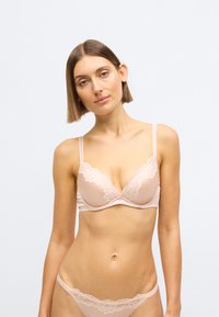 Šviesiai smėlio spalvos apatinių rinkinys su nėriniais, įskaitant gilų liemenėlę su reguliuojamomis petnešėlėmis ir derantį stringų kelnaites, lygaus satino paviršiaus.