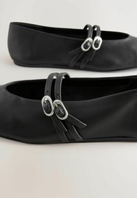 Musta nahkainen ballerinatyyppinen kengät, joissa on pyöreä kärki ja kaksi vastaavaa remmiä sekä hopeiset solkikoristeet päällä yksityiskohtina.