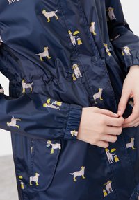 Marin regnrock med en glänsande yta som har ett mönster av hundar som bär gula hattar och stövlar, elastiska manschetter och en justerbar midja.