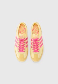 Gule sneakers med rosa snøring og striper. Materialet har en kombinasjon av glatte og semskede teksturer. Logoen er tydelig vist på innsiden.