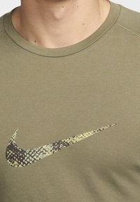 Oliivinvihreä lyhythihainen paita, jossa on pyöreä kaula-auko; siinä on suuri, kankainen Nike-swoosh-logo tummanruskeana ja vaaleankeltaisena.